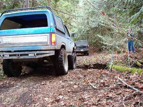 1985 Ford Bronco - A blue Ford Bronco is parked on a dirt path in a wooded area, with a second dark SUV behind it. The Bronco has large off-road tires and a c…
