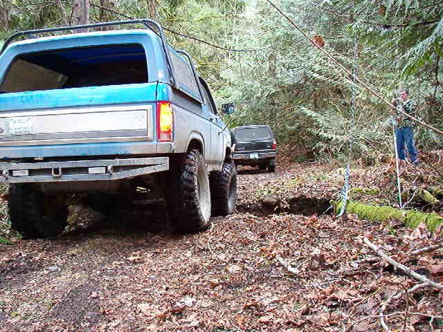 Blue Ford Bronco with off-road tires on a dirt path in a forest.