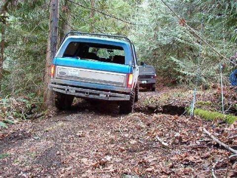 1985 Ford Bronco - A blue Ford Bronco is navigating a muddy, wooded trail, with a second dark SUV visible behind it. The Bronco is angled towards a ditch, wit…