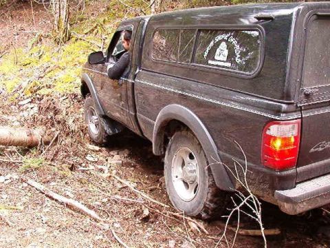 1985 Ford Bronco - A dark-colored pickup truck with a camper shell is driving off-road on a dirt path with trees and brush surrounding it. A sticker with text…