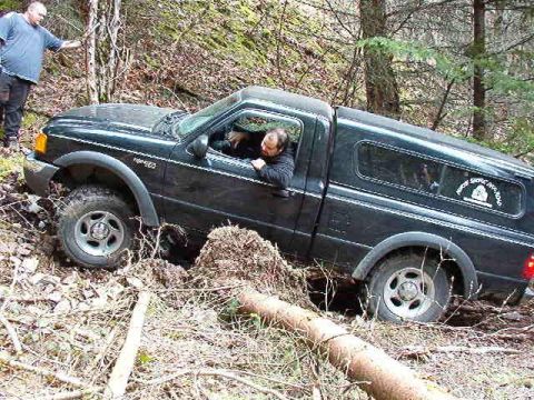 1985 Ford Bronco - A black Ford pickup truck is stuck in a wooded area with a man standing outside and another man driving. The truck is tilted and its front…