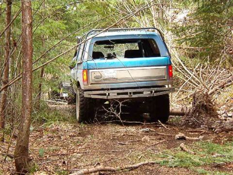 1985 Ford Bronco - The rear view of a blue Ford Bronco is shown driving on a wooded trail. The vehicle has a silver stripe along the side and a custom rear bu…