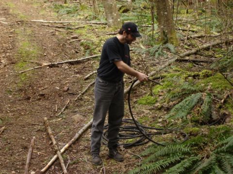 1985 Ford Bronco - A man in a black t-shirt and baseball cap holds a black hose in a wooded area with ferns and mossy ground.