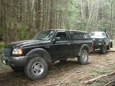 1985 Ford Bronco - A black Ford Ranger pickup truck with a camper shell is parked on a dirt path in a forest, followed by a lifted light blue SUV. Both vehicl…