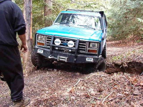 1985 Ford Bronco - A blue Ford Bronco is partially submerged in a ditch off-road, with its front tires stuck in the mud. A person in dark clothing stands to t…