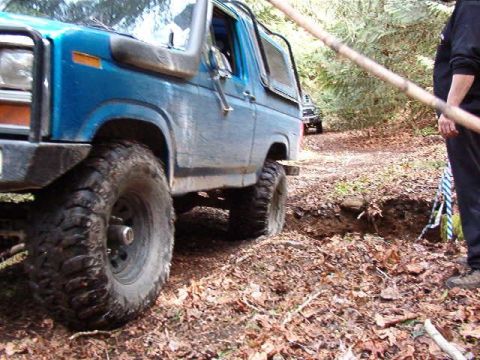 1985 Ford Bronco - A blue Ford Bronco is navigating a rough, wooded trail, with its front tire positioned precariously close to a ditch. The vehicle has large…