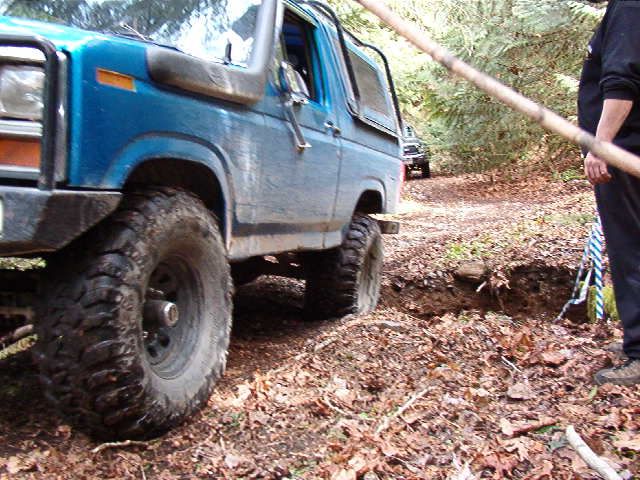 Blue Ford Bronco on a wooded trail with large off-road tires near a ditch.