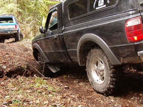 1985 Ford Bronco - A black pickup truck with a camper shell is navigating a rutted dirt path in a wooded area. The front of a blue pickup truck is visible in…