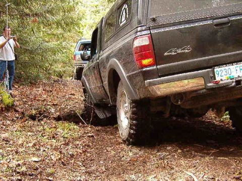 1985 Ford Bronco - A dark-colored pickup truck with a camper shell is driving off-road on a dirt path covered in leaves. Another blue SUV is visible behind it.