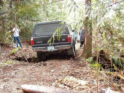 1985 Ford Bronco - A black Ford pickup truck with a camper shell is parked on a dirt path in a wooded area, with two people standing nearby. The truck is angl…