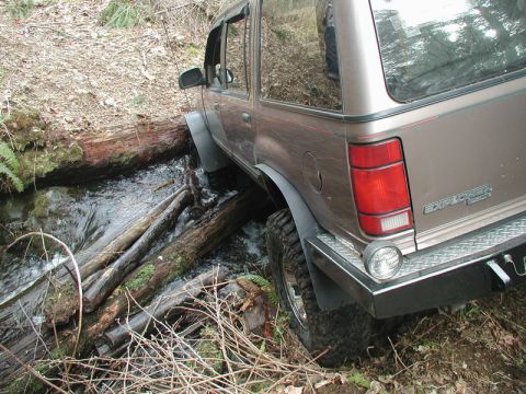 1985 Ford Bronco - A tan Ford Explorer SUV is navigating a stream, with its tires on logs and the vehicle partially submerged.