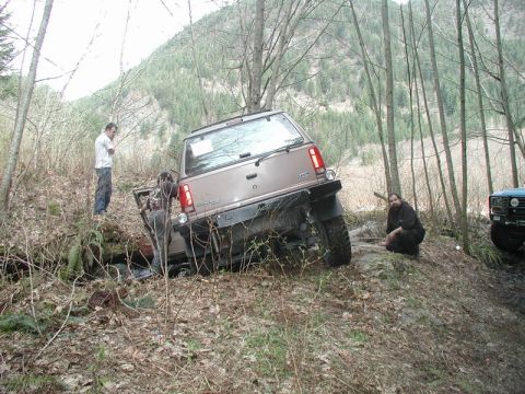 1985 Ford Bronco - A brown Ford Bronco is tilted precariously on a steep, wooded trail, with two people standing nearby and another person crouching on the em…