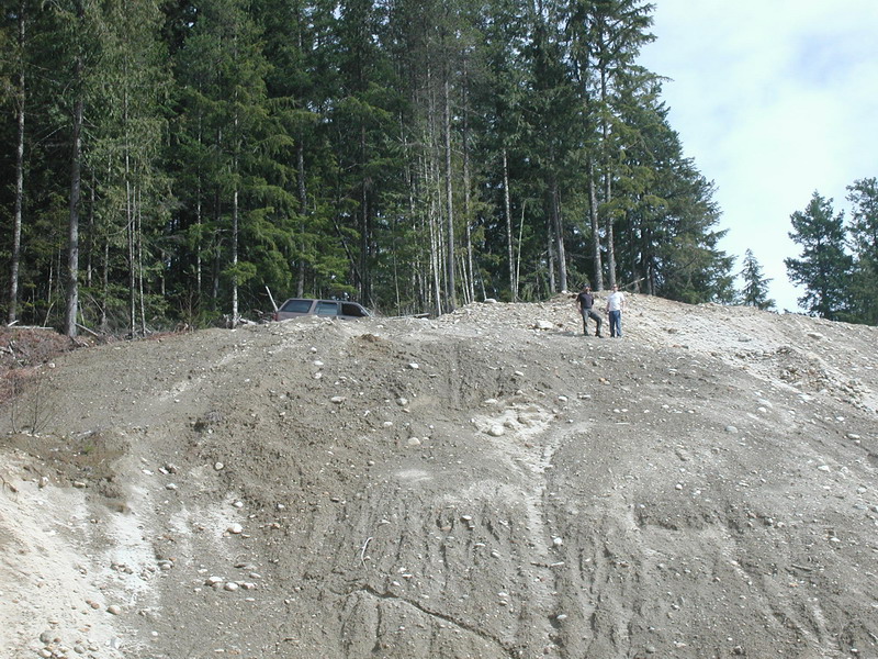 Silver SUV stuck in a dirt hill with two people standing on top of the hill.