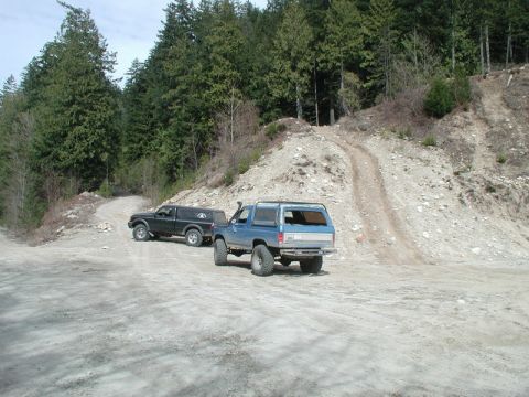 1985 Ford Bronco - Two off-road vehicles are parked on a dirt road in a wooded area. A blue SUV is in the foreground, and a black pickup truck with a camper s…