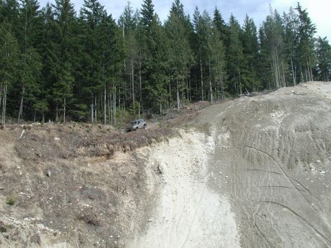 1985 Ford Bronco - A silver SUV is parked precariously on the edge of a steep, eroded hillside, with a dense forest of evergreen trees in the background.