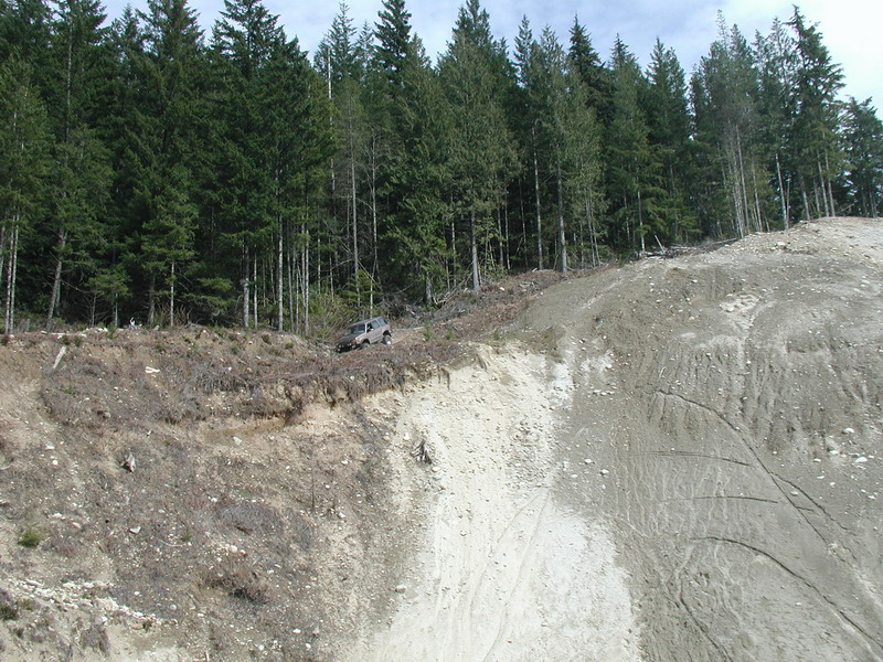 Silver SUV on the edge of a steep, eroded hillside with a forest behind it.