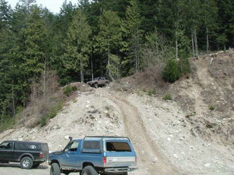 1985 Ford Bronco - Two off-road vehicles are parked on a dirt path in a wooded area. A blue Ford Bronco is in the foreground, and a black pickup truck is to i…