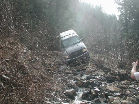 1985 Ford Bronco - A dark-colored SUV has gone off-road and is lodged precariously in a rocky, wooded ravine with a stream running through it. A person is vis…