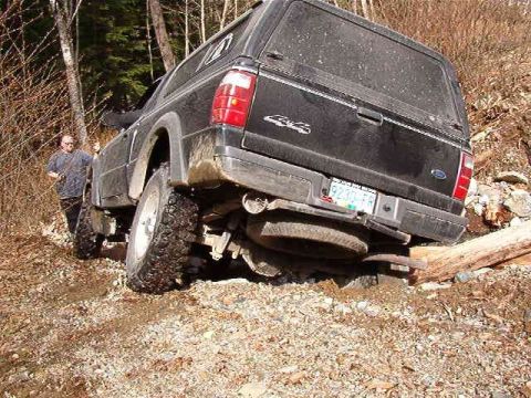 1985 Ford Bronco - A black Ford pickup truck with a camper shell is stuck off-road, with its rear tire resting on a log. A person stands nearby on a gravelly,…