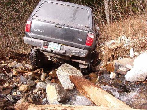 1985 Ford Bronco - The rear of a dark-colored pickup truck, possibly a Ford Bronco, is partially submerged in a rocky stream bed, with its rear tire wedged be…