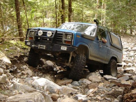 1985 Ford Bronco - A blue Ford Bronco is navigating a rocky, wooded trail, with its front tire lifted high over a boulder. The off-road vehicle features a pro…