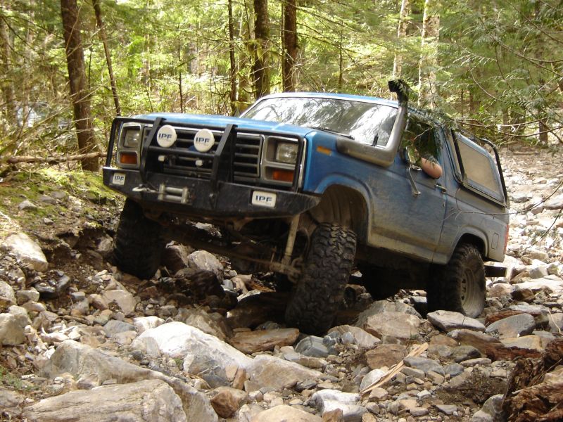 Blue 1985 Ford Bronco off-roading on a rocky trail in a forest.