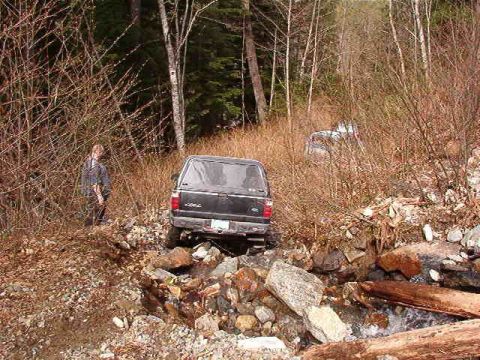 1985 Ford Bronco - A black pickup truck is navigating a rocky, wooded trail. Another vehicle is visible further down the trail.