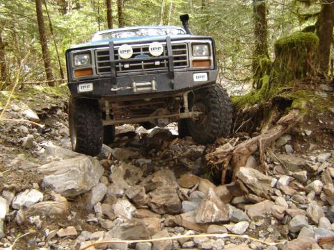1985 Ford Bronco - A blue Ford Bronco is navigating a rocky, wooded trail. The front of the vehicle is angled towards the camera, showing off its off-road tir…