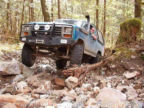 1985 Ford Bronco - A blue 1980s Ford Bronco is navigating a rocky, wooded trail, with its front tires high off the ground. The off-road vehicle features a pro…