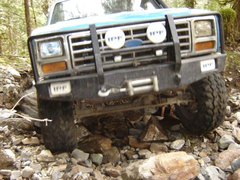 1985 Ford Bronco - The front end of a blue off-road vehicle is shown navigating a rocky terrain with trees in the background. The vehicle has a winch mounted…
