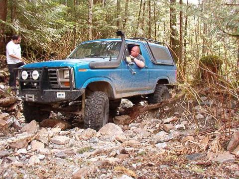 1985 Ford Bronco - A blue Ford Bronco is navigating a rocky, wooded trail. The driver is visible behind the wheel, and another person stands near the front of…