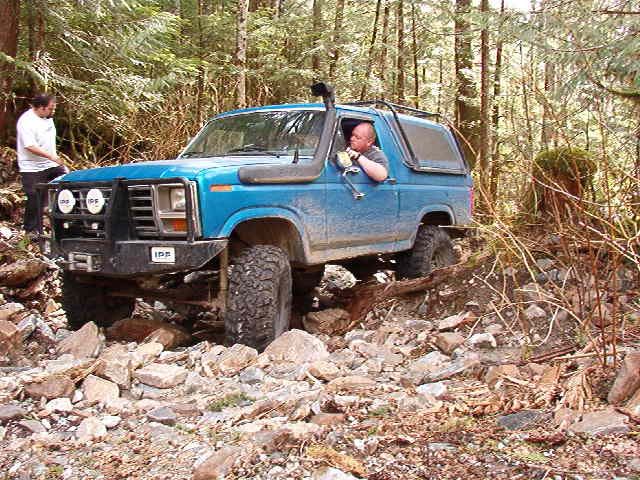 Blue Ford Bronco off-roading on a rocky, wooded trail.