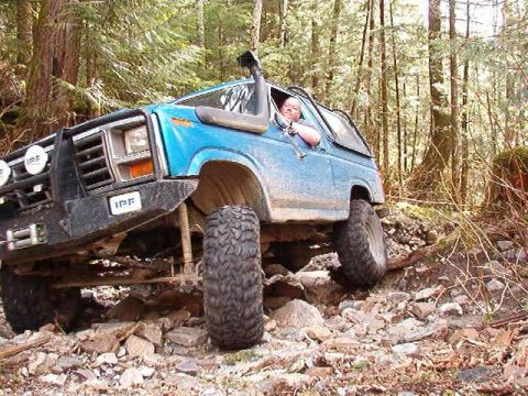 1985 Ford Bronco - A blue 1980s Ford Bronco is navigating a rocky, wooded trail with a person driving. The vehicle is angled to the right, showing its front a…
