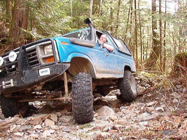 Blue 1980s Ford Bronco off-roading on a rocky trail in a forest.