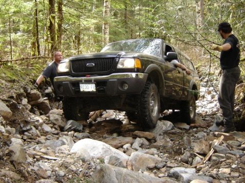 1985 Ford Bronco - A dark-colored Ford pickup truck navigates a rocky, wooded trail with two men present. One man is outside the truck, gesturing, while the o…