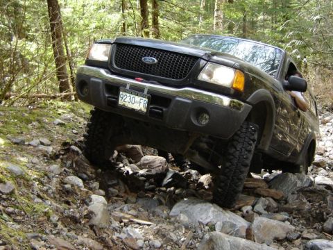 1985 Ford Bronco - A black Ford pickup truck is navigating a rocky, wooded trail with its front tires angled upwards over large stones. The license plate read…