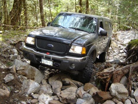 1985 Ford Bronco - A black Ford pickup truck with a camper shell navigates a rocky, wooded trail. The truck's front end is angled towards the viewer, showing…