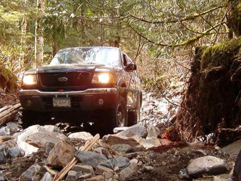 1985 Ford Bronco - A black Ford Ranger pickup truck navigates a rocky, wooded trail. The truck's headlights are on as it drives over a rough terrain.