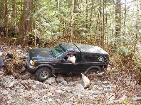 1985 Ford Bronco - A black Ford pickup truck with a camper shell is navigating a rocky, wooded trail. The driver is visible through the open driver's side win…