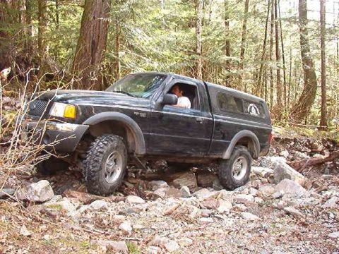 1985 Ford Bronco - A black Ford pickup truck with a camper shell is navigating a rocky, wooded trail. The truck's front tire is angled upwards over a large ro…