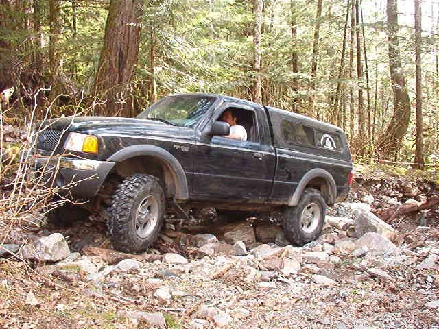 Black Ford pickup truck with camper shell on a rocky, wooded trail.