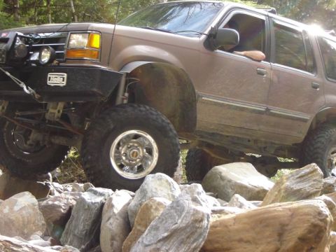 1985 Ford Bronco - A lifted, brown SUV is navigating a rocky terrain, with its front tire high on a boulder. The vehicle has an aftermarket bumper with auxili…