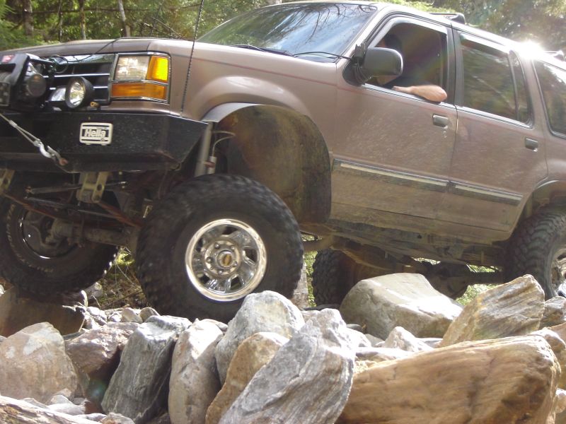 Lifted brown SUV on rocks with aftermarket bumper and lights.