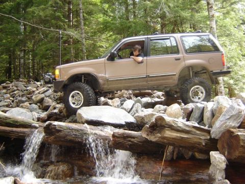 1985 Ford Bronco - A tan Ford Explorer SUV navigates a rocky stream bed, with its front tire high on a rock and its rear tire crossing over a fallen log bridg…