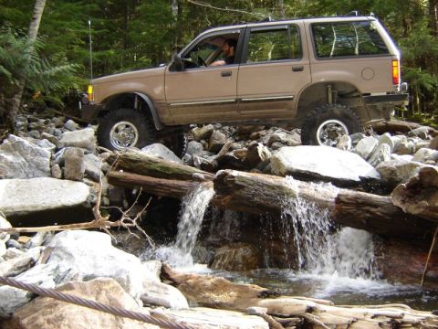 1985 Ford Bronco - A tan Ford Explorer is navigating a rocky stream bed, crossing over fallen logs with water flowing underneath.