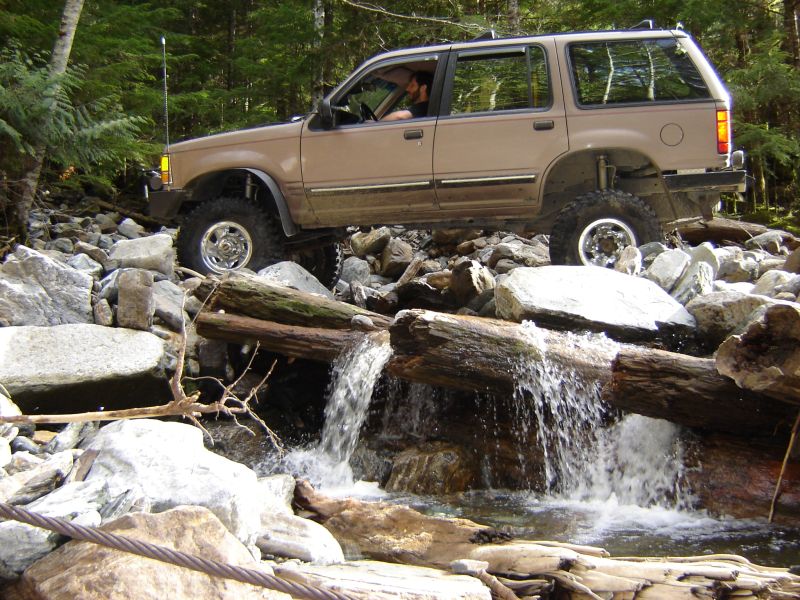 Tan Ford Explorer off-roading over logs and rocks in a stream.