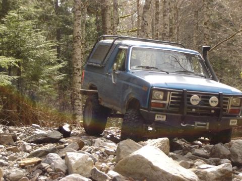 1985 Ford Bronco - A blue Ford Bronco is navigating a rocky, wooded trail. The off-road vehicle features a snorkel, a roof rack, and auxiliary lights on the f…