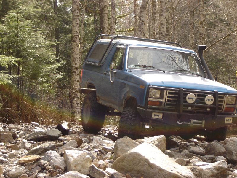 Blue 1980s Ford Bronco off-roading on a rocky trail with trees in the background.