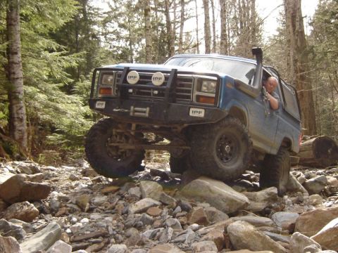 1985 Ford Bronco - A blue 1980s Ford Bronco is navigating a rocky trail in a forest, with its front tire lifted high over a boulder. The vehicle features a sn…