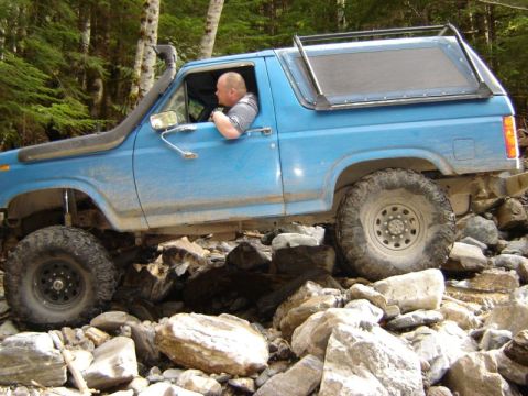 1985 Ford Bronco - A blue Ford Bronco is navigating a rocky, wooded terrain. The driver is visible through the window, and the vehicle is equipped with a snor…